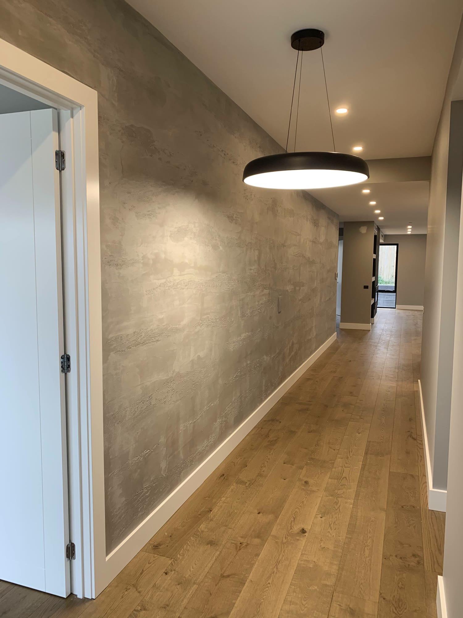 A hallway in bronze-taupe polished plaster running the length of the passage, a round black pendant overhead and oak boards below