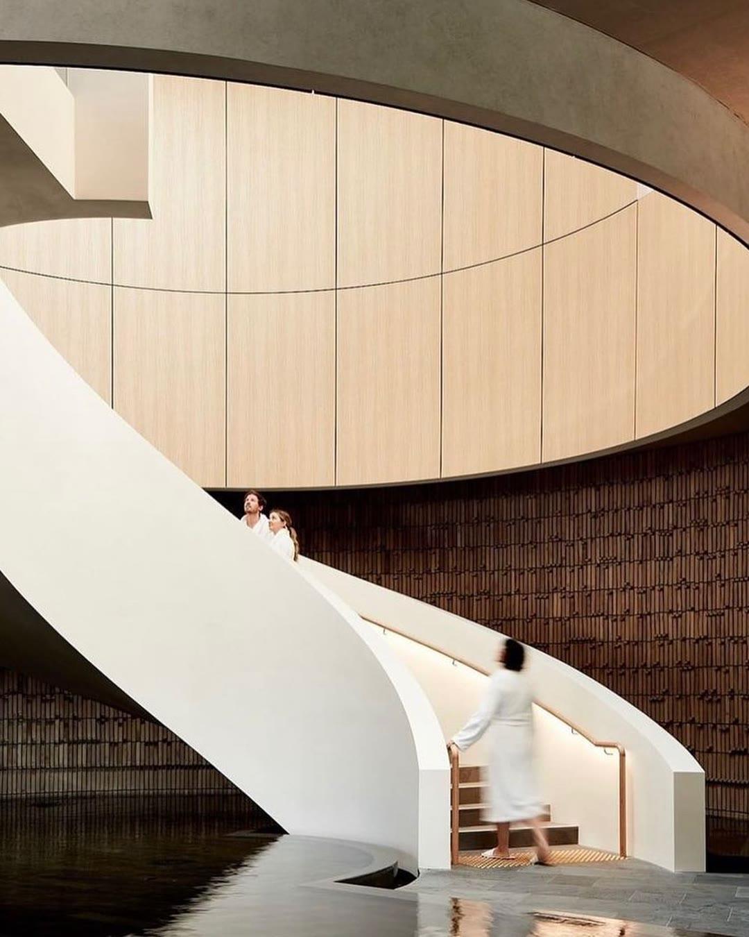 A sculptural white spiral staircase in a double-height space, set against a dark textured wall and a curved timber-lined ceiling