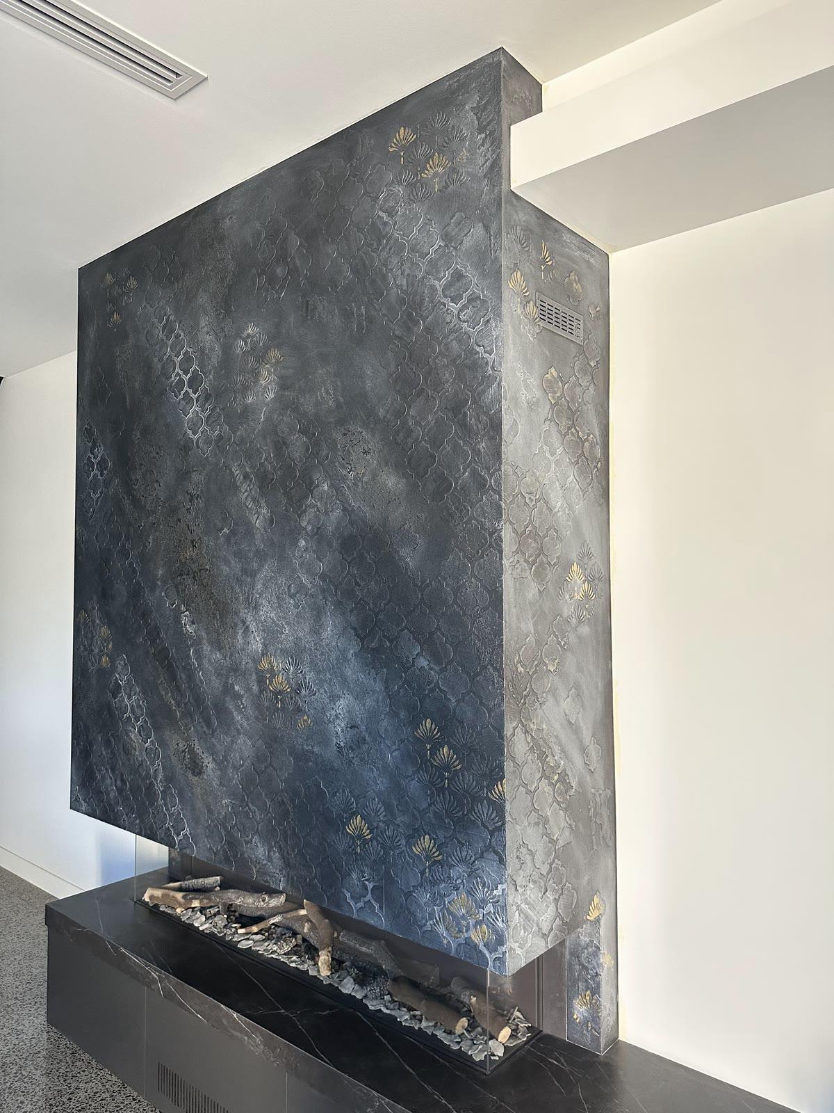 A full-height fireplace breast in charcoal plaster, a scalloped relief pattern worked across it by hand and picked out in gold and silver, standing over a black marble hearth