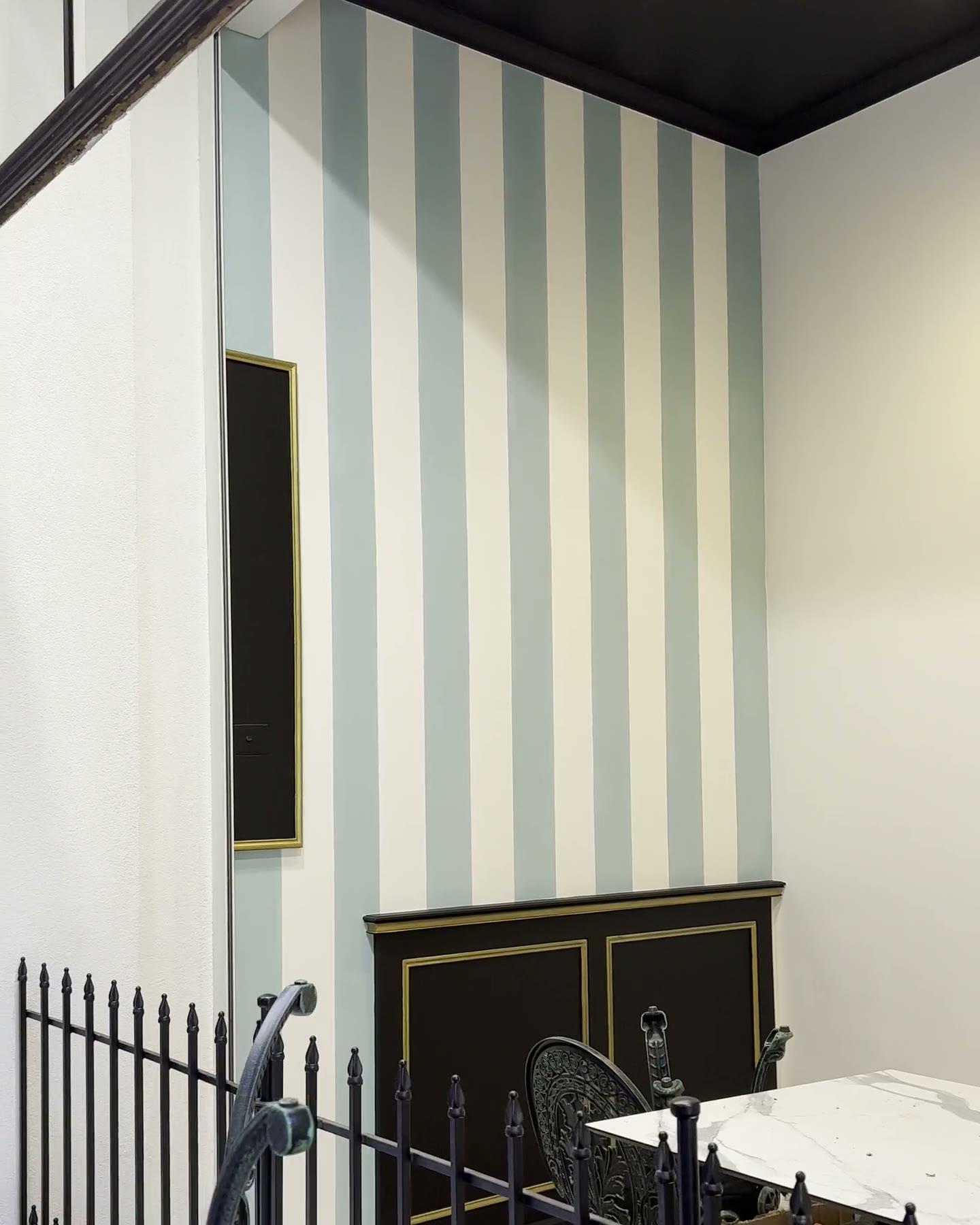 A slow pan across a bistro interior, broad vertical stripes in pale sage and cream running the full height of two walls beneath a black ceiling, above black panelled joinery and a wrought-iron rail