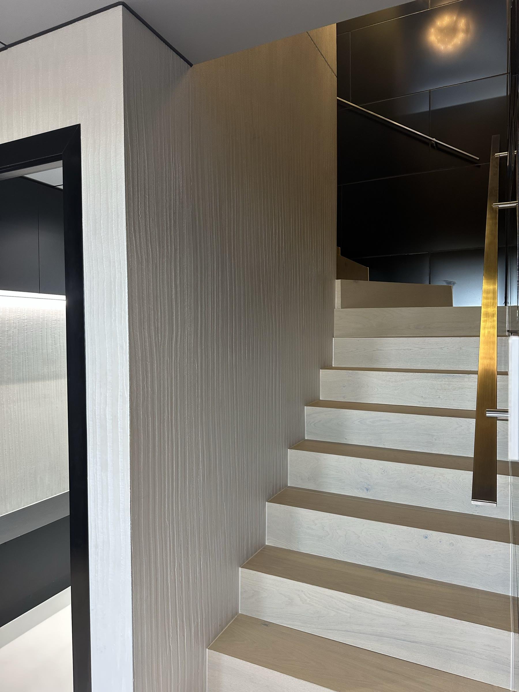 A stairwell wall in pale plaster with fine vertical texture running its full height, beside oak treads and a brass handrail