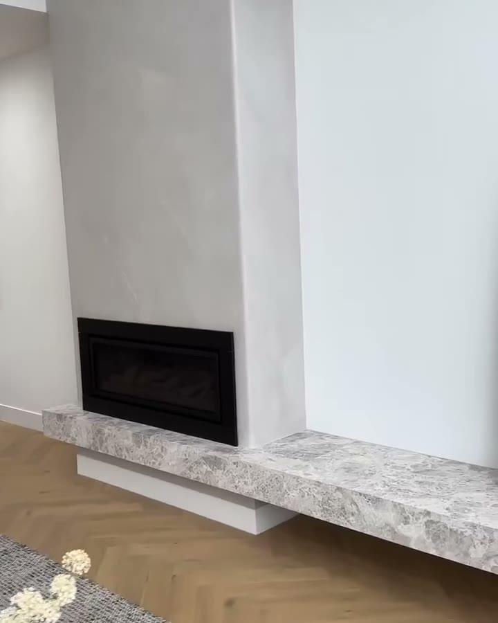 A camera moving past a double-sided fireplace finished in pale silver-grey marbled plaster, a thick stone hearth running out from it over a herringbone timber floor, a dining room visible through the opening