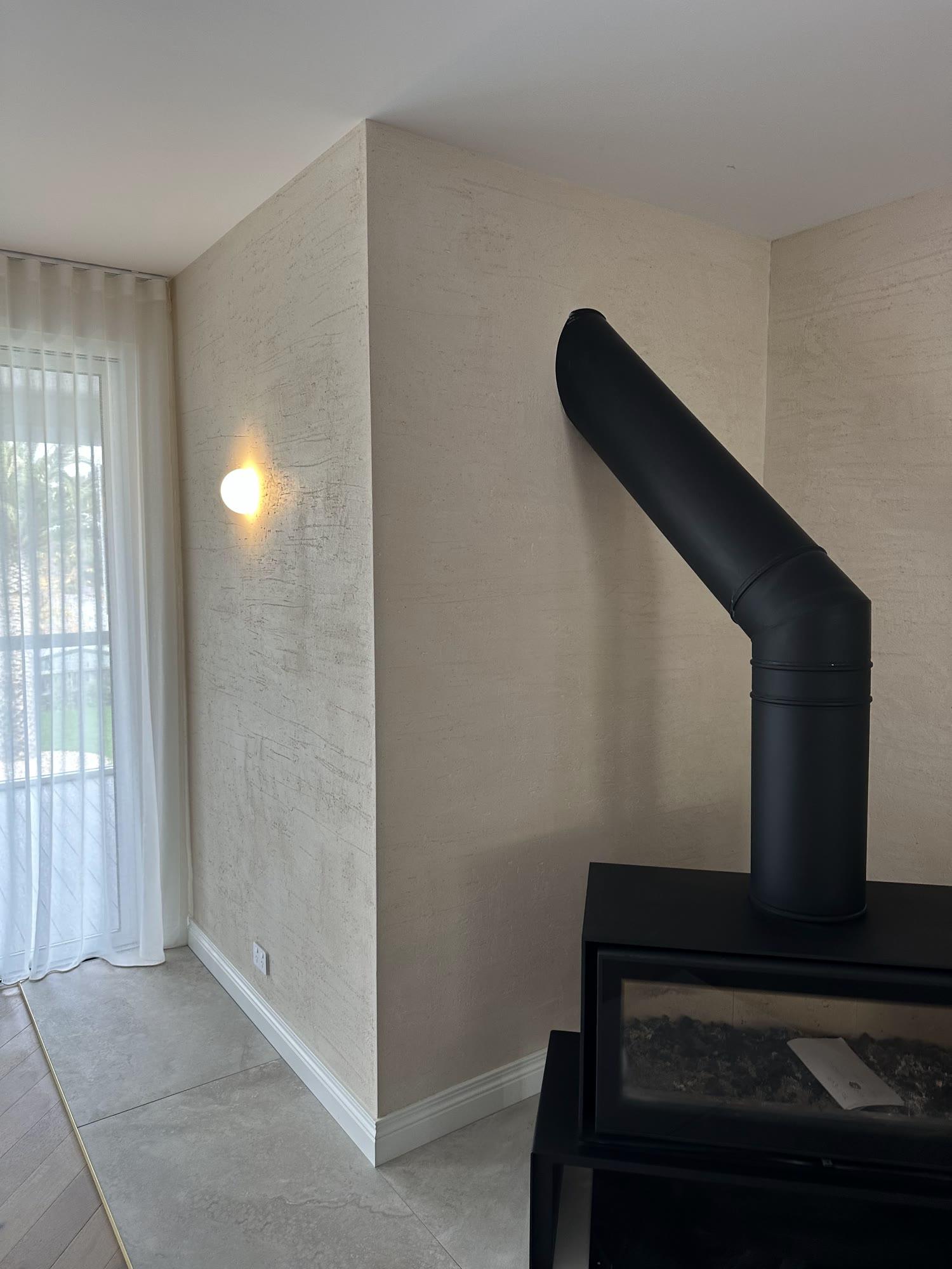 Cream textured plaster running behind a black wood heater and its flue, beside a curtained window